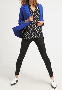 Blazer blu sopra una blusa nera a pois, abbinato a leggings neri e scarpe bianche. L'outfit presenta una silhouette aderente e colori a contrasto.