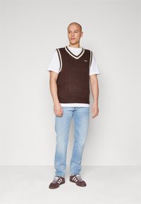 Pulover tricotat, crem, fără mâneci, cu o margine albă în formă de V, purtat peste un tricou alb, asortat cu blugi albaștri deschiși și adidași negri.