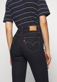 Vaqueros de talle alto azul oscuro con costuras doradas y una etiqueta de cuero en la parte trasera. Presenta un diseño clásico de cinco bolsillos y el logo de Levi's.
