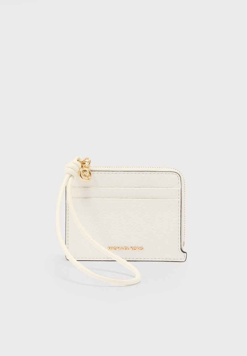 Porte-cartes en cuir blanc avec fermeture éclair dorée, bracelet pour poignet et logo Michael Kors sur la poche avant, sur fond uni.