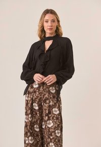 Blusa negra con volantes y escote en forma de ojo de cerradura, combinada con pantalones de pierna ancha marrones con patrones que presentan diseños florales.