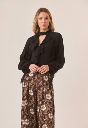 Blusa negra con volantes y escote en forma de ojo de cerradura, combinada con pantalones de pierna ancha marrones con patrones que presentan diseños florales.
