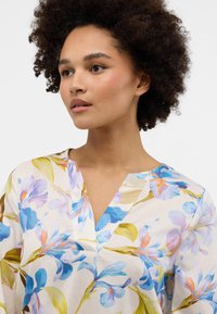 Blusenshirt mit blauen und lila Blumenmustern und grünen Akzenten auf einem cremigen Hintergrund. V-Ausschnitt und leichtes Material.