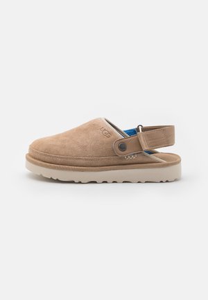 Tan suède instapklompen met een lichte beige rubberen zool, verstelbare achterkant en blauwe textielen voering. UGG-logo aan de zijkant.