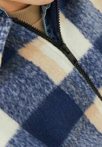 Fleece bunda s modro-krémovým kostkovaným vzorem, se zipem vpředu a jemnou texturou. Přes ní je položena béžová pletená límec.