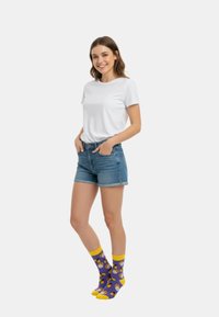 Hvid kortærmet t-shirt, denim shorts og lilla sokker med gule detaljer, prydet med mønstre af bier og blomster. Modellen står med hænderne i lommerne.