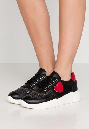 Černé tenisky s třpytivými akcenty, červeným srdcem a záplatami na patě, nošené na holých nohách, s tlustými bílými podrážkami.