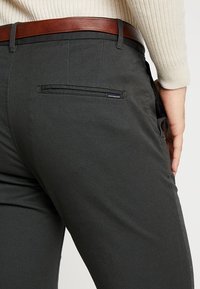 Mörkgrå byxor i slät tyg, med en bakficka med dragkedja, kompletterade med ett brunt skärp i läder.