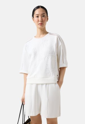 Kvinde iført en hvid kortærmet top med blomsterbroderi og matchende hvide shorts, der holder en taske med sort håndtag.