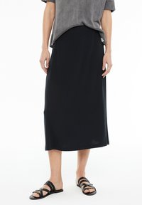 Mujer vestida con una falda midi negra, una camiseta de manga corta gris y sandalias planas negras con tiras trenzadas, de pie sobre un fondo blanco.