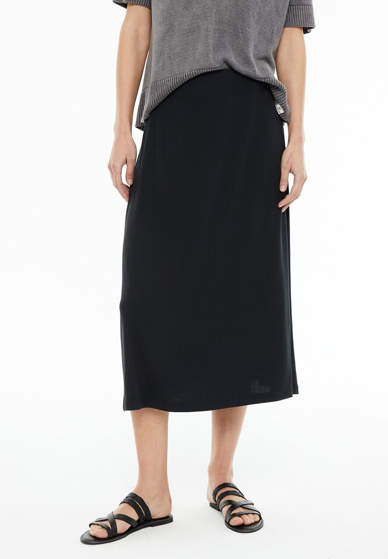 Mujer vestida con una falda midi negra, una camiseta de manga corta gris y sandalias planas negras con tiras trenzadas, de pie sobre un fondo blanco.