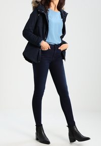 Marineblå polstret jakke med faux fur hætte, lyseblå top, mørke smalle jeans og sorte ankelstøvler med spids snude.