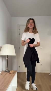 Frau in Freizeitoutfit hält eine schwarze Jacke und steht in einem minimalistischen Raum mit Lampe und Holzboden.