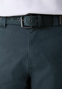 Gros plan sur un pantalon foncé avec une couture frontale et une ceinture en tissu noir tissé attachée par une boucle métallique, portée avec une chemise blanche.