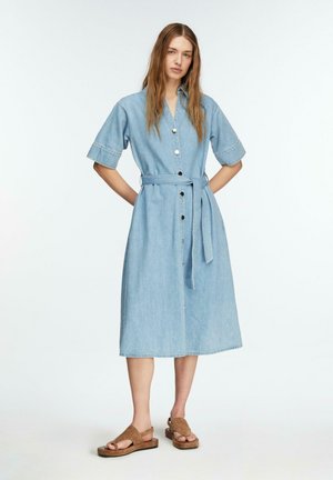 Kobieta ubrana w jasnoniebieską sukienkę midi z denimu z guzikiem z przodu, krótkimi rękawami i paskiem w talii, zestawioną z brązowymi sandałami odsłaniającymi palce.