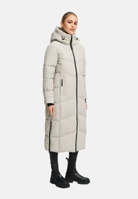 Donna che indossa un lungo cappotto imbottito di un leggero beige con colletto alto e cappuccio, abbinato a pantaloni neri e stivali neri robusti.