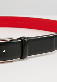 Cinturón de cuero negro con un acabado brillante, con una hebilla de metal plateado y un forro interior rojo. Logo grabado: HUGO HUGO BOSS.