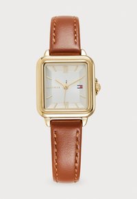 Reloj cuadrado de oro con un dial blanco texturizado, correa de cuero marrón y costuras en contraste de color naranja. Números romanos a las 12 y 6 en punto.