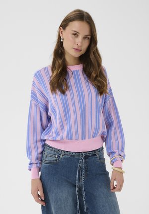 Hullámos hajú nő levendula és kék csíkos pulóvert visel, magas derekú kék farmert kötött övvel, gyöngyfűzár fülbevalót és bézs karkötőket.