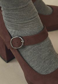 Pantof cu toc înalt din suede maro, prezentând o curea cu cataramă rotundă. Purtați cu șosete texturate gri. Vârf pătrat, toc robust.