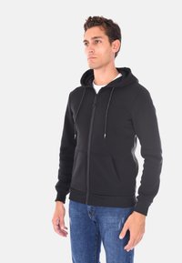 Felpa nera con zip realizzata in tessuto morbido, con cappuccio, cordini, tasche frontali e polsini e orlo a costine. Indossata con jeans blu.