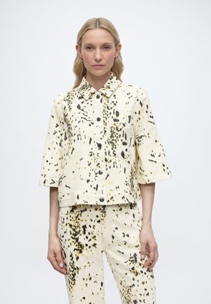 Femme portant une veste et un pantalon crème avec un motif éclaboussé noir et marron, debout devant un fond blanc uni.