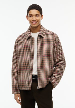 Ung mand smiler iført en beige, brun og rød ternet lynlåsjakke over en hvid skjorte med mørke bukser, hånd i lommen.