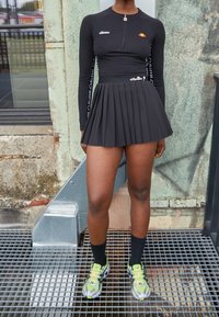 Czarny długi rękaw z zamkiem i logo, zestawiony z plisowaną czarną spódnicą. Noszony z kolorowymi trampkami i czarnymi skarpetkami.