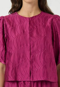 Fuksjowy krótki top z satyny, z teksturowanym wzorem fal, krótkimi bufkami oraz okrągłym dekoltem ze zapięciem na środku przodu.