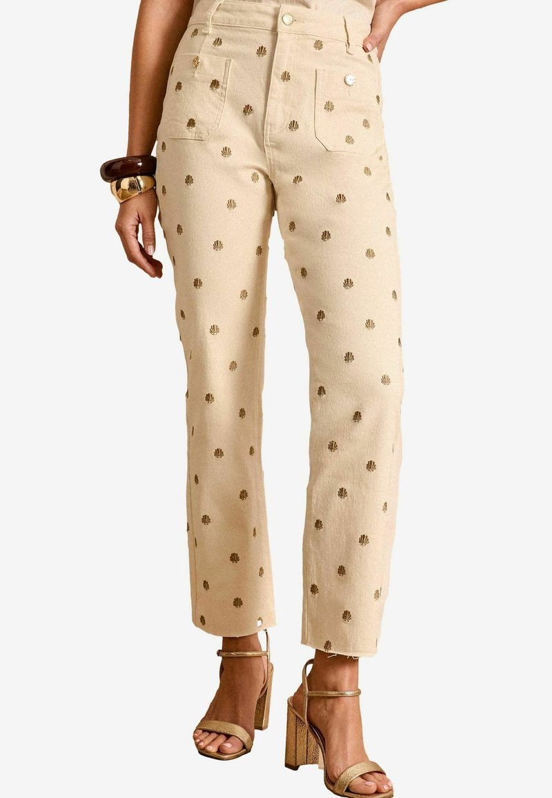 Pantaloni in denim beige con motivi a conchiglia dorati, a vita alta, gamba dritta, con tasche anteriori e posteriori, abbinati a tacchi alti con cinturini.
