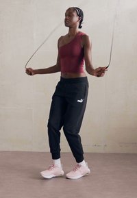 Vrouw met gevlochten haar die binnen touwtjespringt, gekleed in een bordeauxrood tanktop, zwarte Puma-sweatpants en roze sneakers.