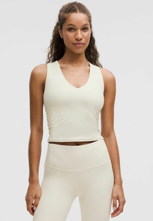 Woman with curly hair wearing a sleeveless, cream-colored ribbed top and matching high-waisted leggings, standing against a plain background.
