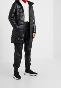 Persoon in een zwarte pufferjack, zwarte joggingbroek, wit shirt met een rode coltrui eronder, en zwarte instap Sneakers.