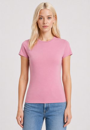 Młoda kobieta z długimi blond włosami stoi przodem, ubrana w prosty różowy t-shirt z krótkim rękawem i niebieskie jeansy na jasnym tle.