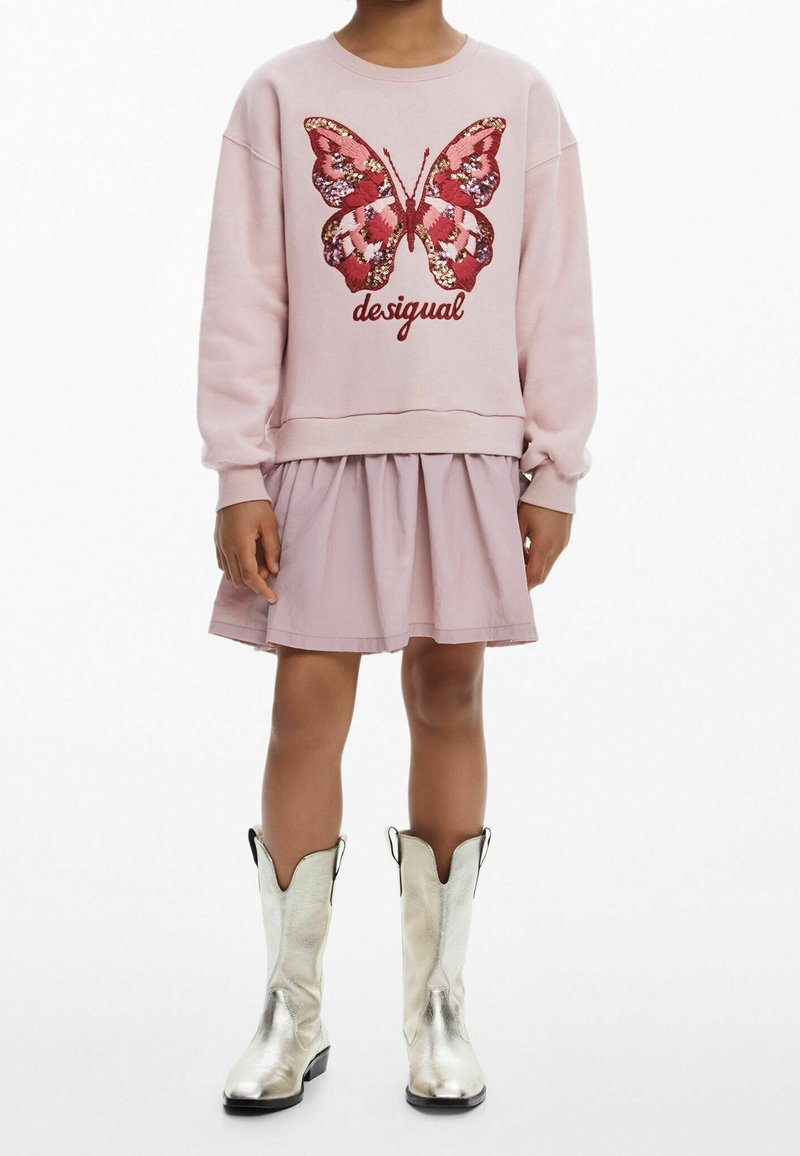 Enfant portant une robe rose clair et un sweat-shirt avec un grand papillon rose et rouge et le mot «desigual», assorti à des bottes argentées brillantes.
