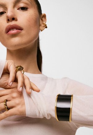 Femme portant des boucles d'oreilles créoles en or, plusieurs bagues en or et un large bracelet noir et or sur un haut léger à manches longues, posant les mains près du visage.