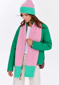 Ručne pletená ružovo-zelená šatka s rebrovanými koncami, nosená so zelenou bundou. Mäkká textúra a žiarivý farebný kontrast. Detaily zahŕňajú dekoratívny prívesok.