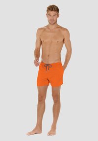 Oranssit uimapöhikät, joissa on nyöri vyötäröllä, tarjoavat sileän tekstuurin ja yksinkertaisen muotoilun. Pöhikät päättyvät reiteen puoliväliin, ilman kuvioita tai lisäosia.