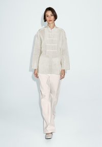 Camicetta lungo maniche color beige chiaro, con collo e strisce verticali, chiusura anteriore con bottoni, abbinata a pantaloni larghi color crema.
