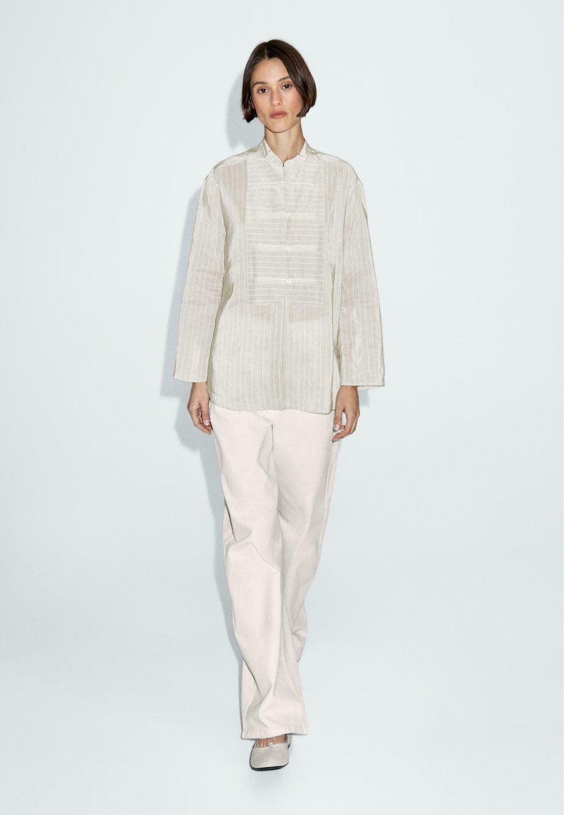 Camicetta lungo maniche color beige chiaro, con collo e strisce verticali, chiusura anteriore con bottoni, abbinata a pantaloni larghi color crema.