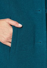 Türkiser Wollmantel mit sichtbaren Nähten, in dem eine Hand in einer Tasche steckt. Der Stoff hat eine weiche Textur, mit einer geknöpften Vorderseite und einem maßgeschneiderten Design.