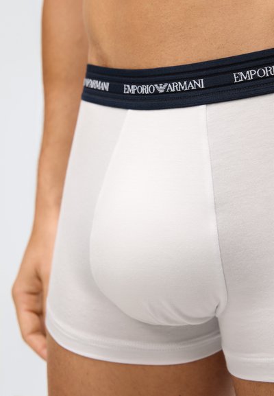 Detailní záběr osoby nosící bílé trenýrky Emporio Armani s černým páskem a viditelným logem na jednobarevném pozadí.