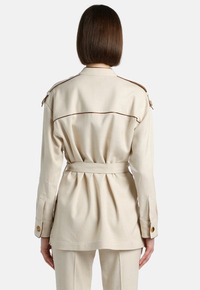 Beige belted takki, jossa on laskostettu selkäosa, ruskea kanttaus olkapäällä ja nappi-mansetit. Sileä pinta ja muotoiltu malli.
