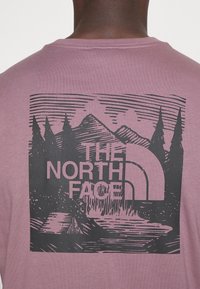 Fialové bavlněné tričko s černým grafickým designem hor, stromů a vodopádu, s textem "THE NORTH FACE".