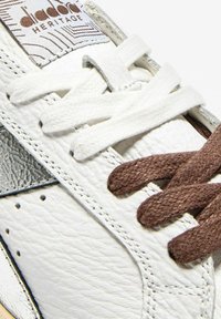 Sneaker in pelle bianca con superficie texture, pannello grigio e lacci a contrasto marroni e bianchi. Etichetta Diadora Heritage visibile.
