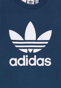 Mörkblå bomulls t-shirt med en stor vit Adidas-logotyp med tre ränder och en treflor-design centrerad ovanför varumärkesnamnet.