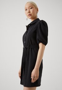 Robe en denim noir avec un col, des manches bouffantes, un devant boutonné et une taille ceinturée. Le mannequin porte des boucles d'oreilles en forme de cœur et des bagues.