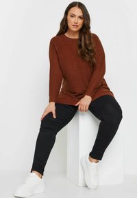 Rostfärgad ribbad tröja, svarta slim-fit byxor och vita sneakers. Modellen sitter på en vit pelare, med vågigt hår som hänger ner.