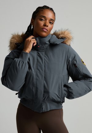 Mujer con chaqueta acolchada gris oscuro con capucha forrada de piel, cerrándola con cremallera, sobre un fondo claro y liso.