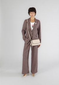 Ljust brunt linnekostym med vida byxor och skräddarsydd kavaj. Vit topp med färgglad läppbrodering. Beige axelväska med sömdetaljer.